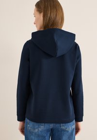 Sweat à capuche bleu marine avec une poche kangourou, des poignets côtelés et une capuche à cordon. Texture douce, coupe ajustée, porté avec un jean bleu clair.
