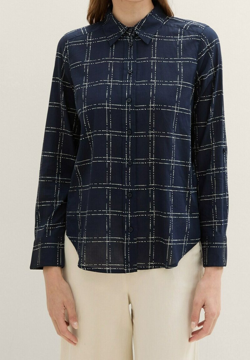 Femme portant une chemise bleu marine à manches longues avec un motif à carreaux en fenêtre blancs en pointillé et un pantalon beige.