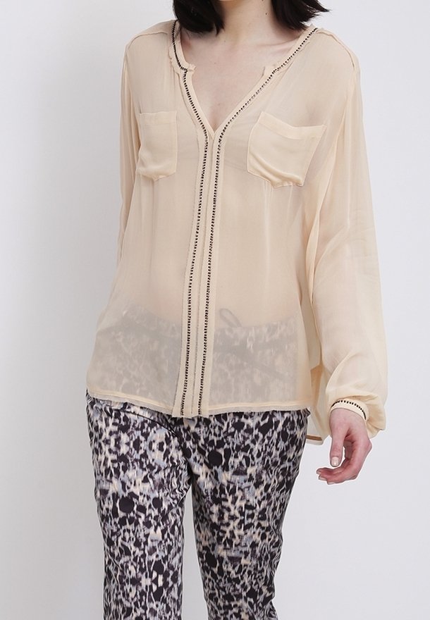 Femme portant un chemisier beige transparent avec un fin liseré noir et deux poches poitrine, associé à un pantalon à motifs noir et blanc.