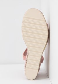 Semelle d'une sandale plate beige clair avec des rainures horizontales texturées, présentée en position verticale contre un fond d'angle blanc.