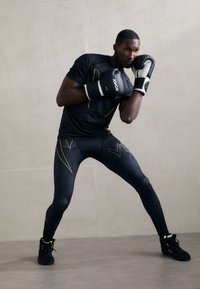 Hombre con ropa atlética negra y guantes de boxeo en una posición defensiva de boxeo frente a un fondo de pared lisa.
