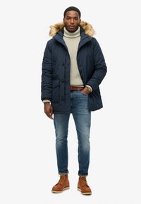 Parka blu navy con cappuccio bordato in pelliccia, chiusura con bottoni, tasche laterali, abbinata a un dolcevita crema, jeans blu e stivali marroni.