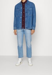 Man i blå denimjacka och jeans, bordeauxfärgad skjorta, vita sneakers, stående mot en enfärgad vit bakgrund.