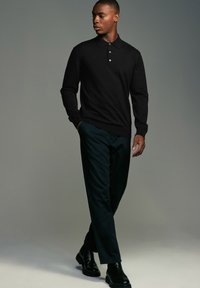 Melnā polo krekla ar garām piedurknēm, apkakli un trim pogām, apvienots ar tumši zaļām biksēm un melniem potīšu zābakiem. Gluda auduma tekstūra.