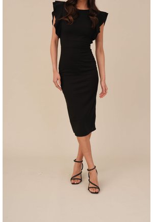 Kvinde iført en tætsiddende sort midi-kjole med flæser på korte ærmer og sorte remsandaletter med høje hæle, stående på en ensfarvet beige baggrund.