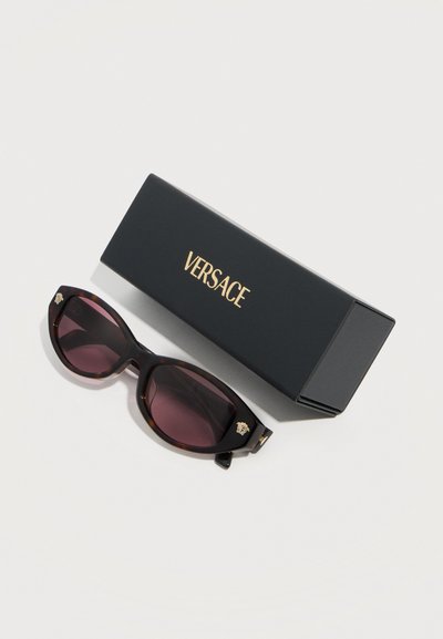 Μαύρα γυαλιά ηλίου cat-eye με μοβ φακούς και χρυσό λογότυπο Medusa, τοποθετημένα δίπλα σε μια μαύρη θήκη με την επωνυμία Versace πάνω σε λευκή επιφάνεια.