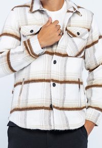 Rutig skjortjacka med fleecefoder i vitt med bruna ränder, med två bröstfickor, svarta knappar och klassisk krage.