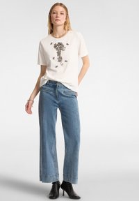Camiseta blanca de manga corta con adornos florales plateados, combinada con jeans de pierna ancha en azul claro y botas de tacón negras hasta el tobillo.