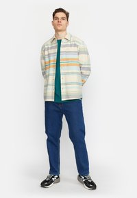 Licht gekleurde gestreepte button-up shirt over een turquoise shirt, gecombineerd met blauwe jeans en zwarte sneakers met witte accenten.