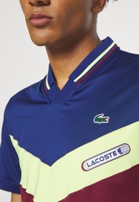 Kortärmad polotröja med färgblock i blått, gult och vinrött. Har en v-ringning, horisontella ränder och Lacoste-logotyp på bröstet.