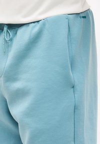 Κοντινό πλάνο ανοιχτό γαλάζιου φούτερ παντελονιού με ελαστική μέση και κορδόνι, φορεμένου με λευκό τοπ, που δείχνει πλαϊνή τσέπη και υφή υφάσματος.