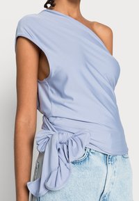 Vrouw in een lichtpaarse one-shoulder top met een grote strikdetails aan de zijkant, gecombineerd met lichtblauwe denim jeans.