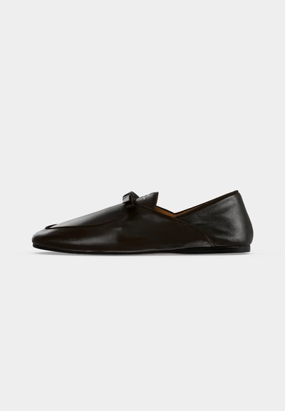 Μαύρο δερμάτινο μοκασίνι slip-on με μικρή λουρίδα στο πάνω μέρος, εμφανίζεται σε πλαϊνή όψη σε απλό φόντο.
