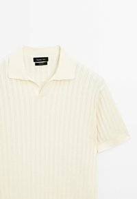 Polo-shirt en tricot à manches courtes de couleur crème avec une texture à côtes verticales et un col doux, disposé à plat sur un fond blanc.