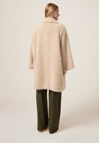 Béžový chlupatý kabát s výstřihovými límci, širokými rukávy a jemnou texturou. V kombinaci s tmavě zelenými, volně střiženými kalhotami.