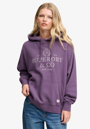 Ung kvinde med langt blond hår iført en lilla Superdry & Co hættetrøje og blå jeans, med hænder delvist i lommerne.