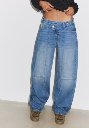Personne portant un jean bleu ample avec un double bouton à la taille et des baskets marron, levant un t-shirt noir pour montrer le ventre.