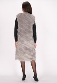 Grau kunstpelz Weste mit langem, weichem Material; taillierte Form; gestufter Look; getragen über einem schwarzen Langarmshirt, vor einem schlichten Hintergrund präsentiert.
