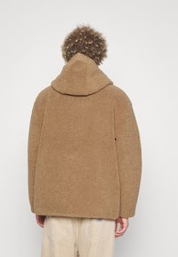 Sudadera de felpa marrón con una superficie texturada, capucha y corte relajado. Presenta un diseño sin costuras y se lleva con pantalones de pana de color claro.