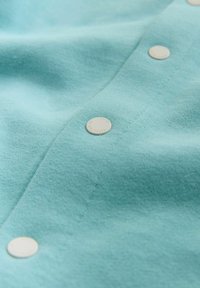 Turkusowa tkanina o gładkiej fakturze z okrągłymi srebrnymi guzikami wzdłuż zapięcia, podkreślająca minimalistyczny styl i konstrukcję.