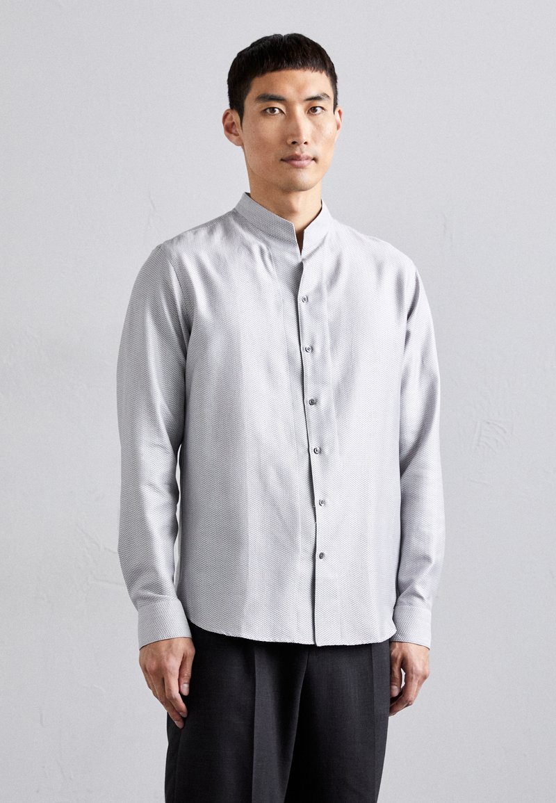 Camisa de botón de color gris claro con cuello alzado y textura estampada. Mangas largas y un acabado de dobladillo sutil, combinado con pantalones oscuros.