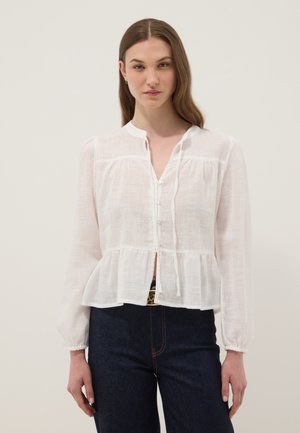 Mujer con blusa blanca de manga larga con botones y bajo peplum, y vaqueros oscuros de talle alto, sobre un fondo liso.