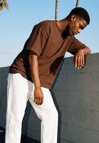 Barna inges és fehér nadrágos férfi, aki egy szürke betonfalnak dől kültéren, háttérben pálmafákkal és kék éggel.