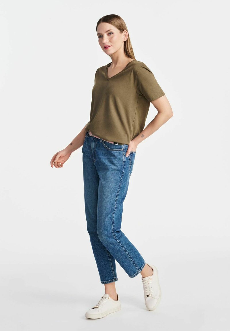 Oliwkowy, krótki rękaw, bluzka typu V połączona z jasnoniebieskimi dżinsami o tapered cut i białymi trampkami, zaprezentowana na neutralnym tle.