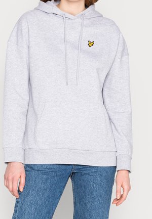 Kobieta ubrana w jasnoszary kaptur z małym żółtym emblematem ptaka na piersi oraz w niebieskie dżinsy.