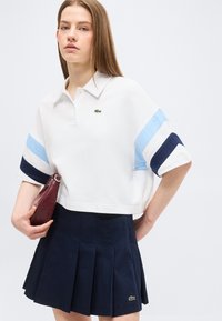 Νεαρή γυναίκα που φοράει λευκή κοντομάνικη πόλο με μπλε ρίγες και σκούρο μπλε πιε ντε πουλ φούστα, κρατώντας μια μικρή μπουρντό τσάντα χειρός.