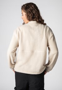 Fleecejacke in Creme mit strukturierter Oberfläche, hohem Kragen und überschnittenen Schultern. Mit Naht im oberen Rückenbereich und Seitentaschen.