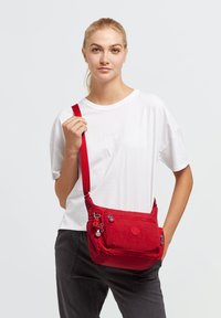 Red shoulder bag made of textured fabric, featuring multiple zippered compartments and a sturdy adjustable strap. Round logo detail on front.