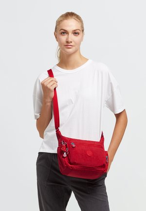 Red shoulder bag made of textured fabric, featuring multiple zippered compartments and a sturdy adjustable strap. Round logo detail on front.