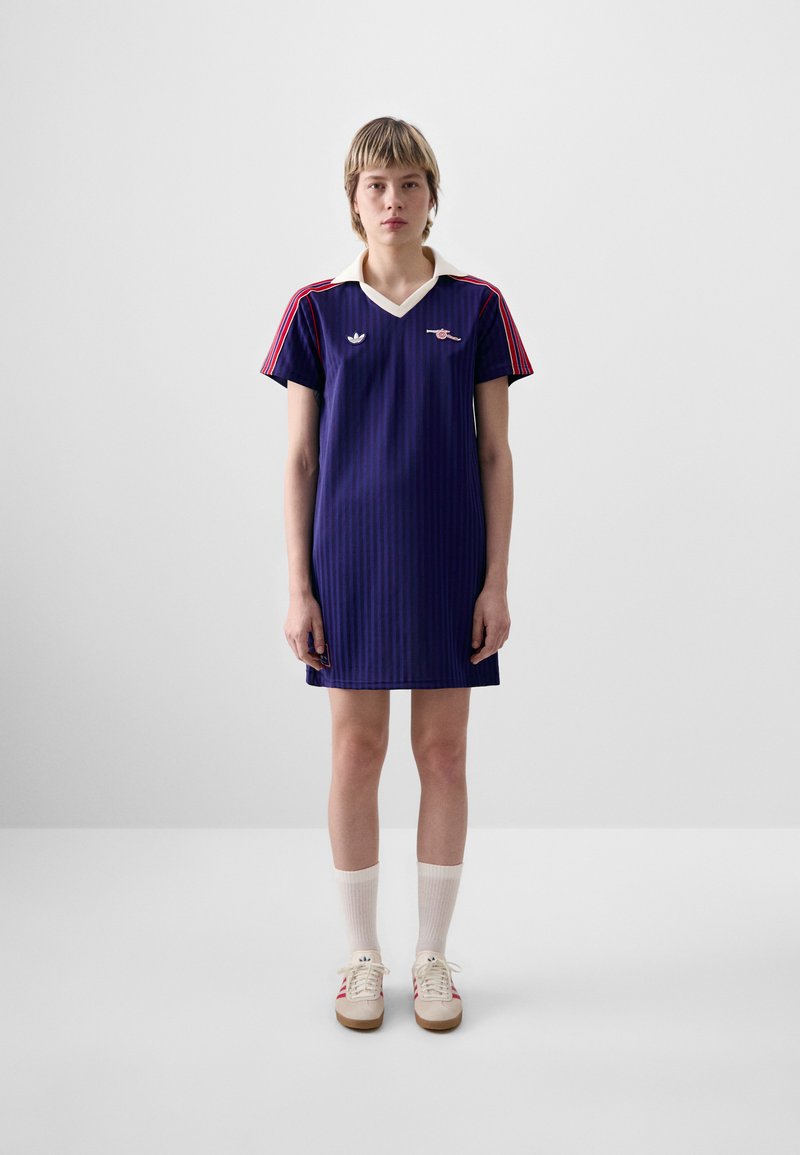 Robe rayée bleu marine à manches courtes, col blanc et accents rouges. Portée avec des chaussettes blanches et des chaussures beiges ornées de rayures rouges.