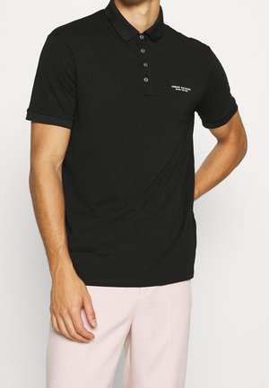 Man draagt een zwarte Armani Exchange polo met korte mouwen en vier knopen, en lichtroze broek, voor een effen achtergrond.