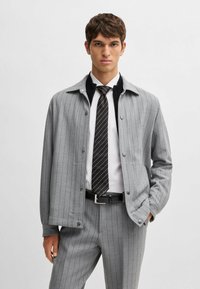 Veste grise à rayures avec des boutons, comportant un col noir. Associée à une chemise blanche et une cravate noire. Design ajusté, tissu texturé.