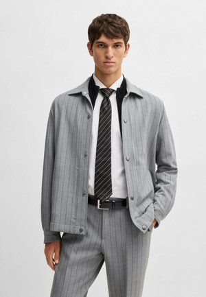Veste grise à rayures avec des boutons, comportant un col noir. Associée à une chemise blanche et une cravate noire. Design ajusté, tissu texturé.