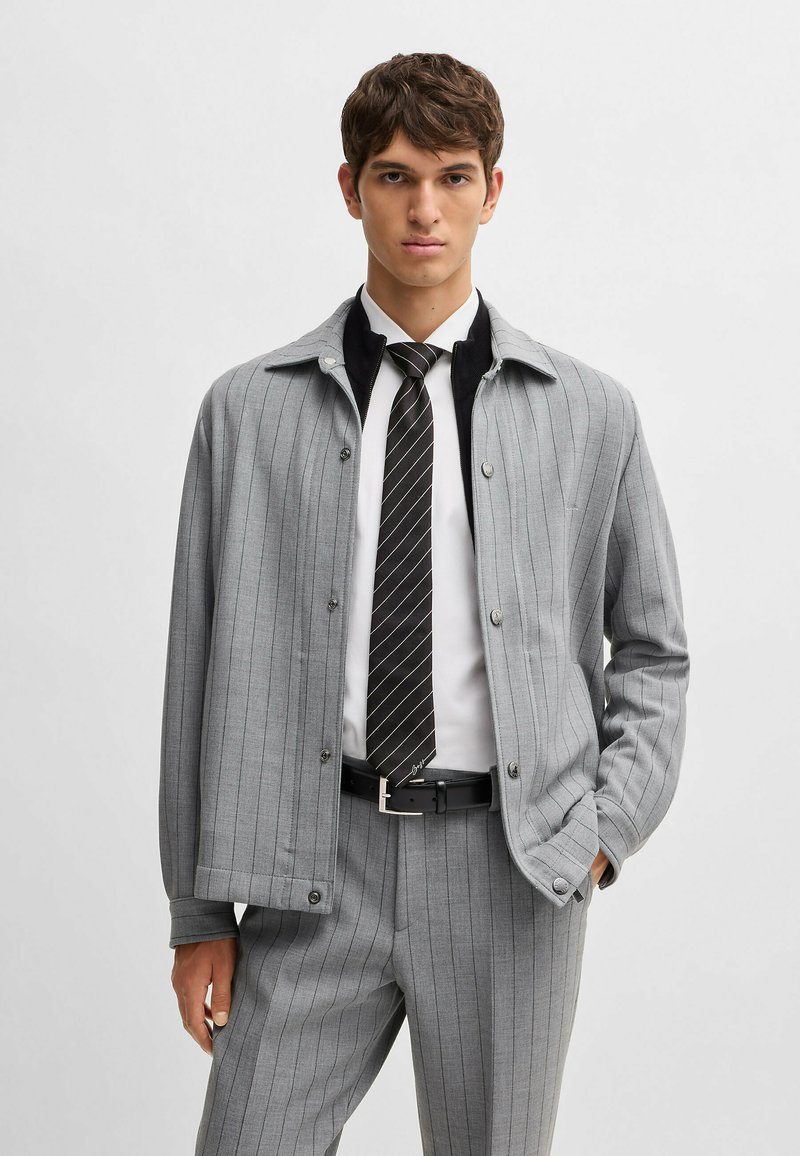 Veste grise à rayures avec des boutons, comportant un col noir. Associée à une chemise blanche et une cravate noire. Design ajusté, tissu texturé.