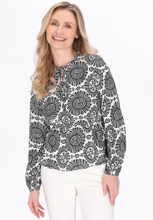Uśmiechnięta kobieta w czarno-białej bluzce z długim rękawem, wzorzystej, z wiązaniem przy szyi, ubrana w białe spodnie, na białym tle.