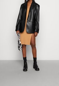 Svart läderblazer över en ribbad, kamelfärgad klänning med slits i sidan. Svarta klackstövlar och en mönstrad väska fulländar looken.