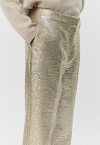 Auksinės, tekstūruotos metalinės plačios kelnės su subtiliu raštu, turinčios juosmenį, kišenes ir lygų paviršių.