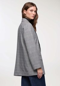 Giovane donna con un blazer a quadri grigi e pantaloni larghi neri, che guarda oltre la spalla su uno sfondo semplice e chiaro.