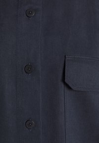 Chemise à boutons bleu marine en tissu texturé, avec une poche et des boutons noirs ornés d'un simple détail de couture.