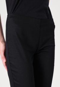 Svarte tettsittende bukser laget av et glatt, elastisk stoff. Har høy midje og detaljer langs sidesømmen. Ingen synlig maskinvare eller mønstre.