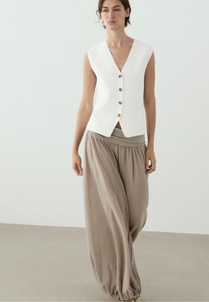 Donna che indossa un gilet bianco smanicato con bottoni e pantaloni beige larghi, in piedi su uno sfondo chiaro e semplice.