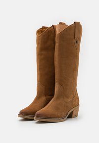 Botas de ante marrón hasta la rodilla con punta afilada, tacón apilado y costuras decorativas a lo largo del borde superior y los lados.