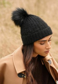 Caciula din tricot neagră cu un model texturat și un pompon din blană sintetică în vârf. Purtați de o persoană cu părul lung, asortată cu o haină de culoare maro deschis.