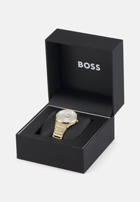 Reloj de pulsera dorado con un dial redondo blanco y pulsera en tono dorado, presentado en una caja negra con el logo "BOSS" en el interior.