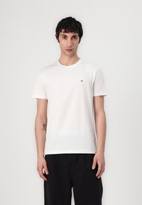Tricou alb din bumbac, cu mâneci scurte, decolteu rotund și manșete răsucite. Prezintă un logo mic pe piept. Stilizat cu pantaloni negri.