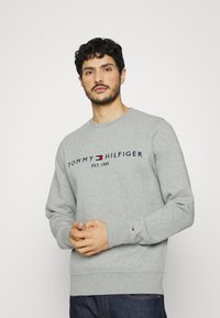 Sudadera gris hecha de tela suave, con cuello redondo, mangas largas y un logotipo con acentos rojos y azules en el pecho.